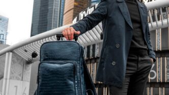 Mann mit Koffer und Rucksack steht vor einer Skyline und wartet auf die Abreise